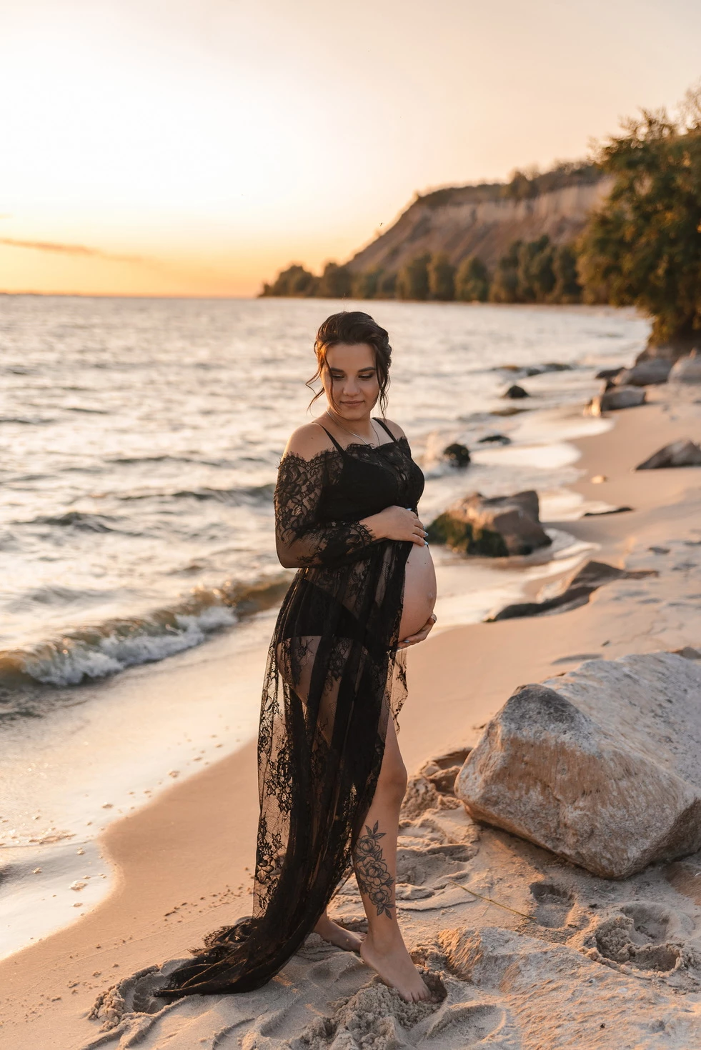 Вагітна жінка стоїть на пляжі в чорному мереживному платті, дивлячись на захід сонця.