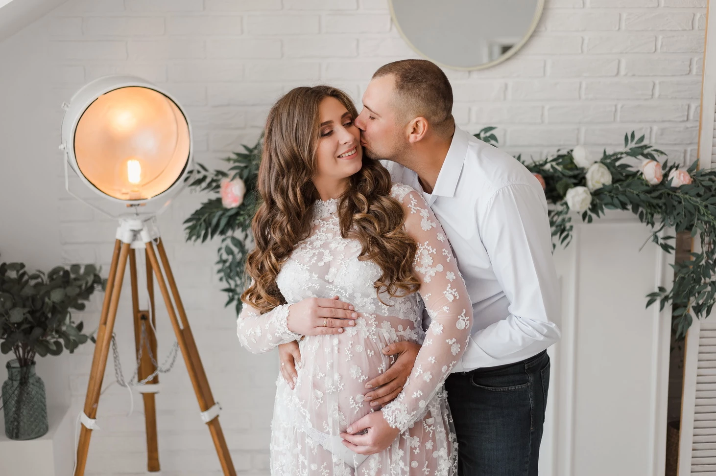 Фотосесія вагітності в Кременчуці: пара в білому вбранні в фотостудії, серед великих ваз і рослин, обіймаються і цілуються.