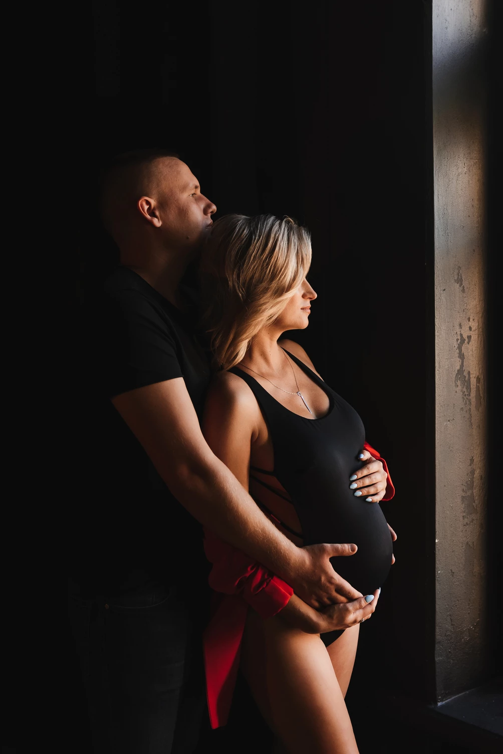 Чоловік обіймає вагітну дружину стоячи біля вікна у фотостудії, де м'яке природне світло ніжно підкреслює їхні обличчя та емоційну близькість.