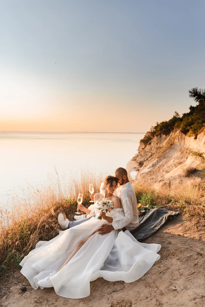 Весільне фото з парою, що обіймається та тримає келихи на фоні моря, відображаючи літній настрій та романтичні моменти на відпочинку.