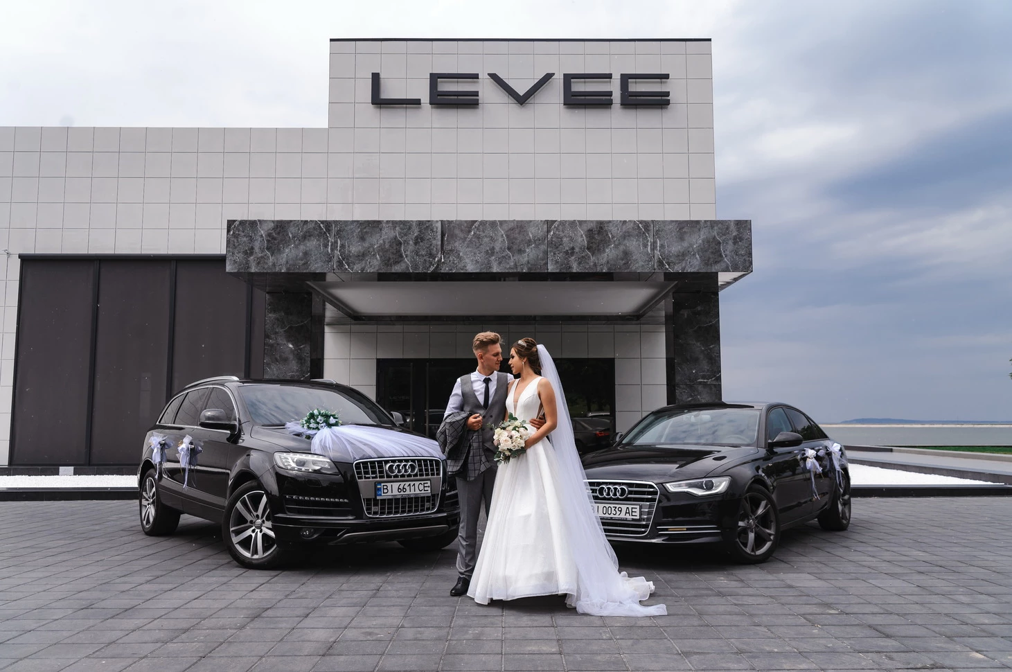 Закохана пара у весільних вбраннях позує на фотографії між двома шикарними Audi, з рестораном Levee, місцем святкування, на задньому плані.