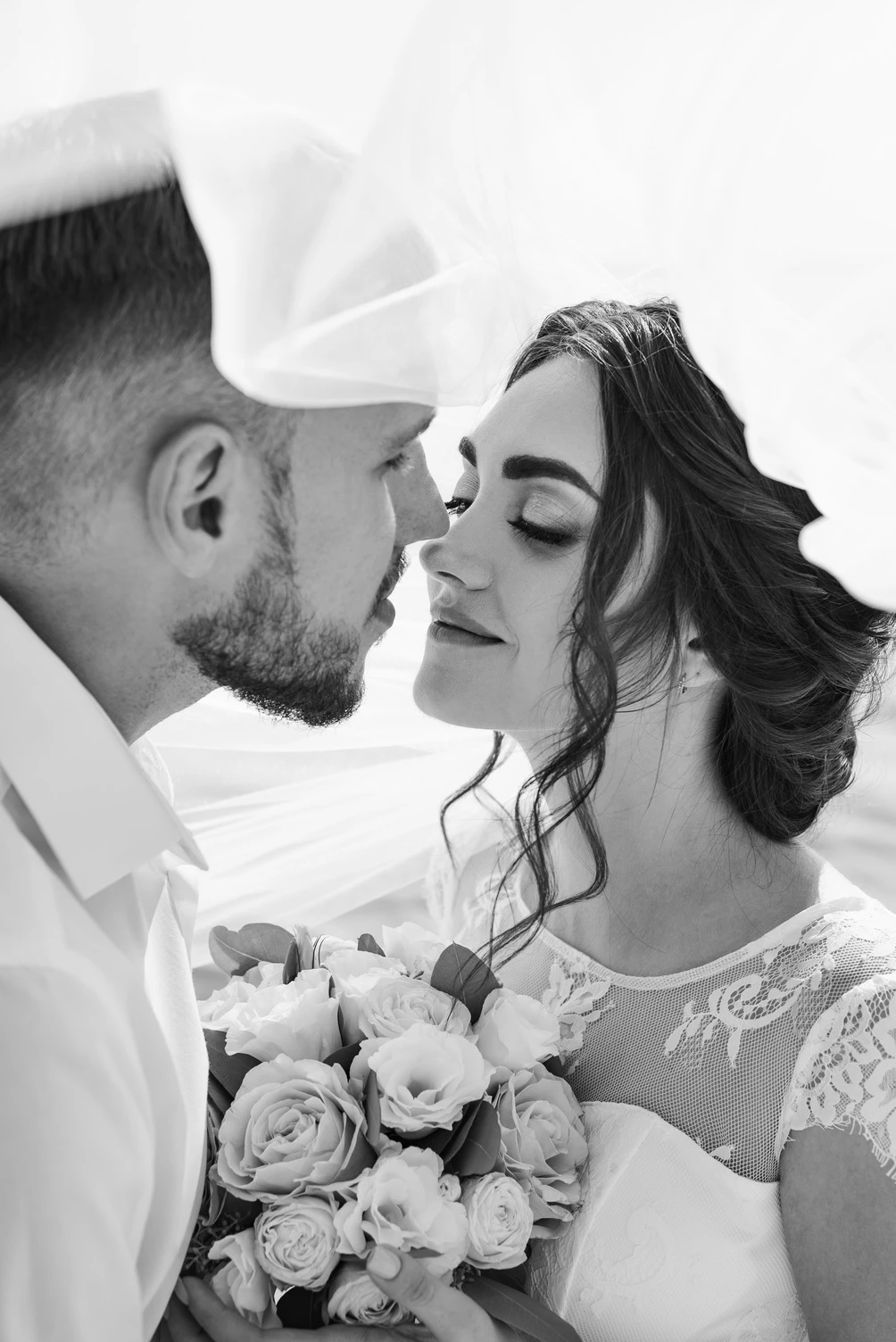 Крупний план чорнобілої весільної фотографії, де наречені обмінюються ніжним поцілунком, з прекрасним букетом квітів між ними.